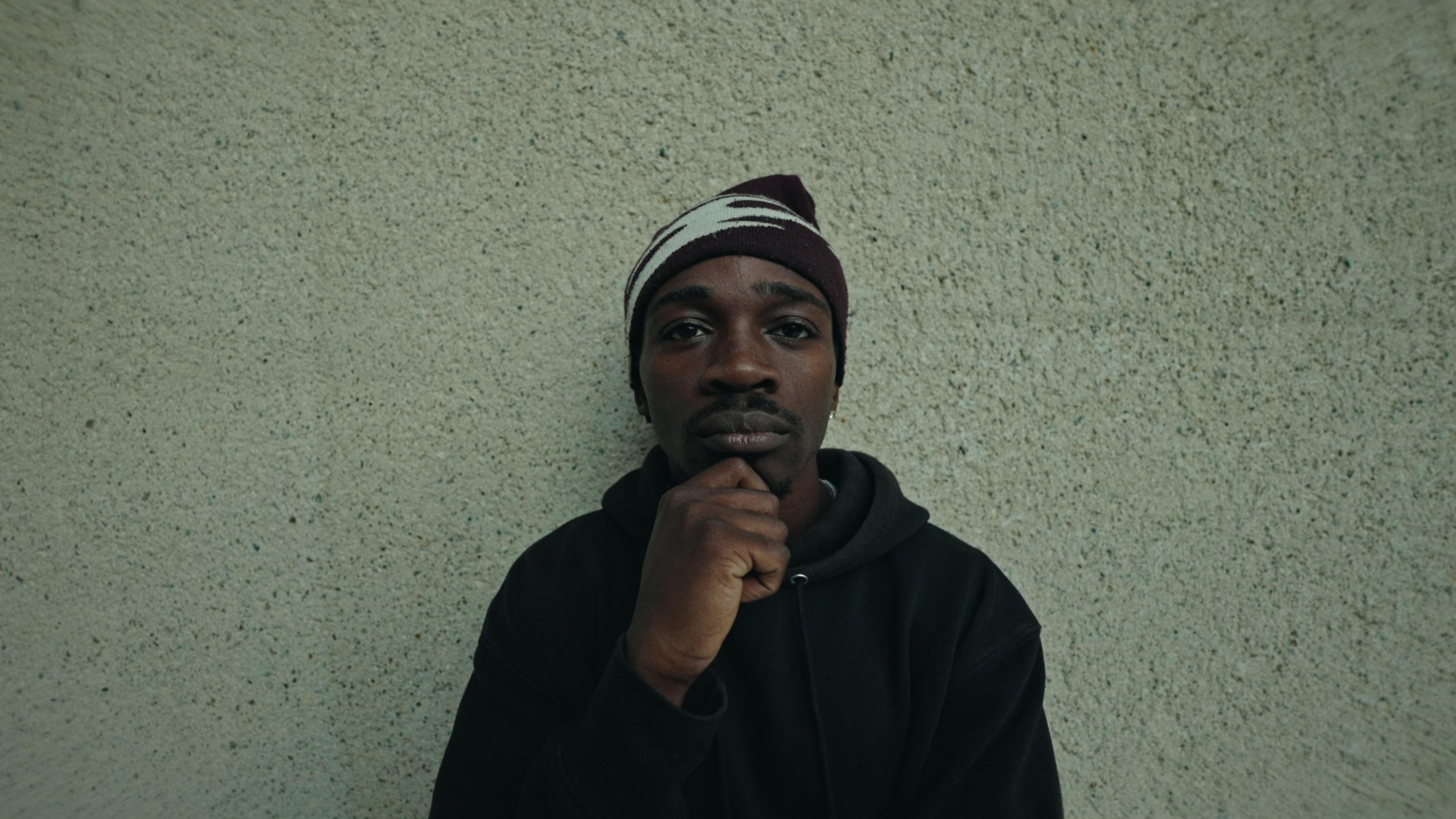 A dancer in a beanie seated against a stone wall.