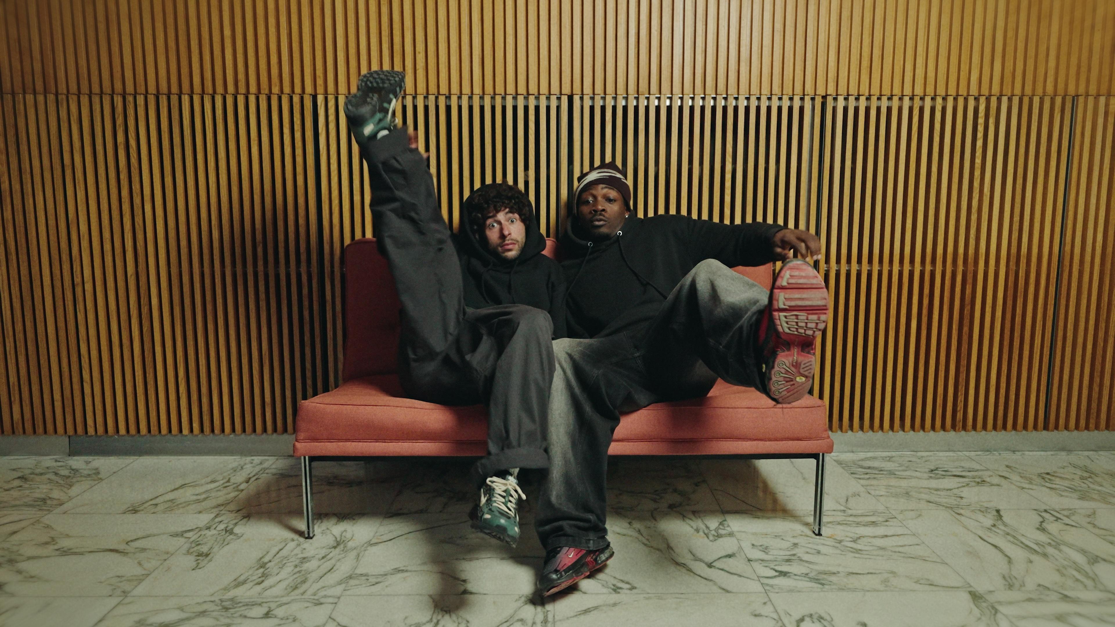 Two dancers sharing an orange chair in front of a wooden slat wall.